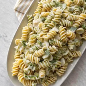Large white platter of rotini pasta salad with creamy pesto sauce and fresh herbs on a marble surface.
