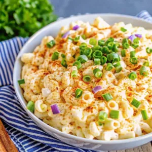 A side view of a large white bowl filled with creamy macaroni salad topped with sliced green onions and dusted with paprika.