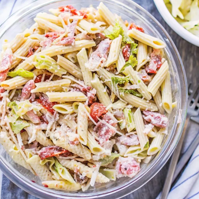 Large glass bowl of penne pasta salad mixed with sun-dried tomatoes, parmesan cheese, and leafy greens.