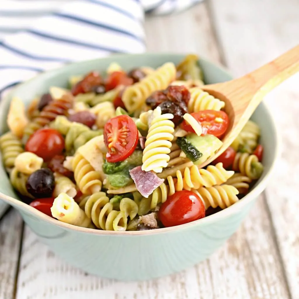 A wooden serving spoon scooping up tricolor pasta salad with tomatoes, cucumbers, and olives from a light blue bowl on a white wooden table.