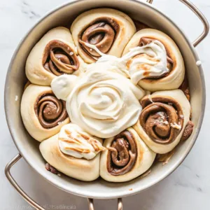 Pull-apart cinnamon rolls arranged in a circular flower pattern in a silver pan with a large swirl of white frosting in the center.