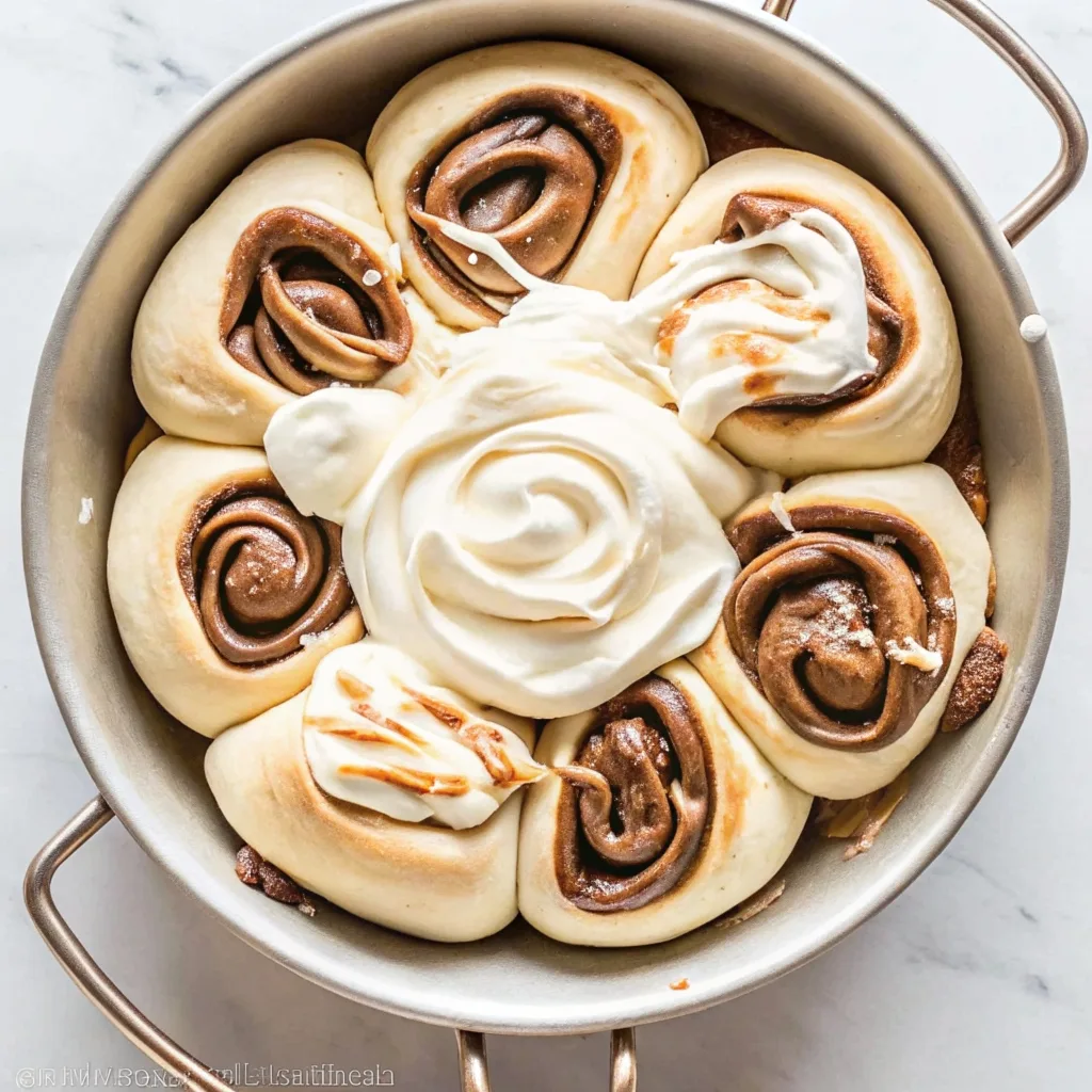 Pull-apart cinnamon rolls arranged in a circular flower pattern in a silver pan with a large swirl of white frosting in the center.