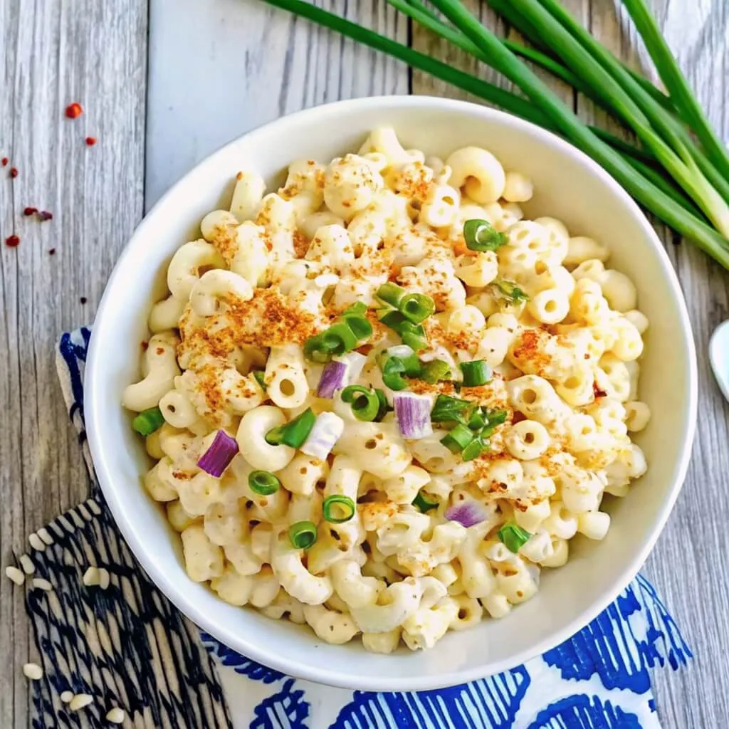 Top-down view of a white bowl containing macaroni salad garnished with green and purple onions on a light wooden background.