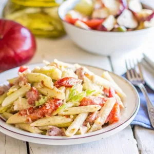 A single serving of penne pasta salad on a white plate with a background featuring a red onion and a secondary salad bowl.