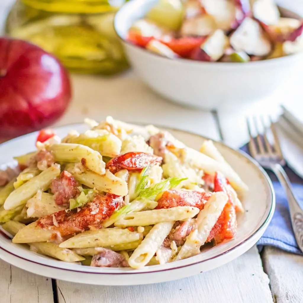 A single serving of penne pasta salad on a white plate with a background featuring a red onion and a secondary salad bowl.