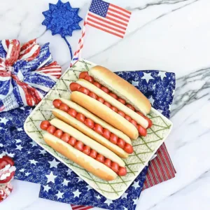 Four hot dogs topped with red spherical garnishes on a square plate, surrounded by American flag-themed decor.