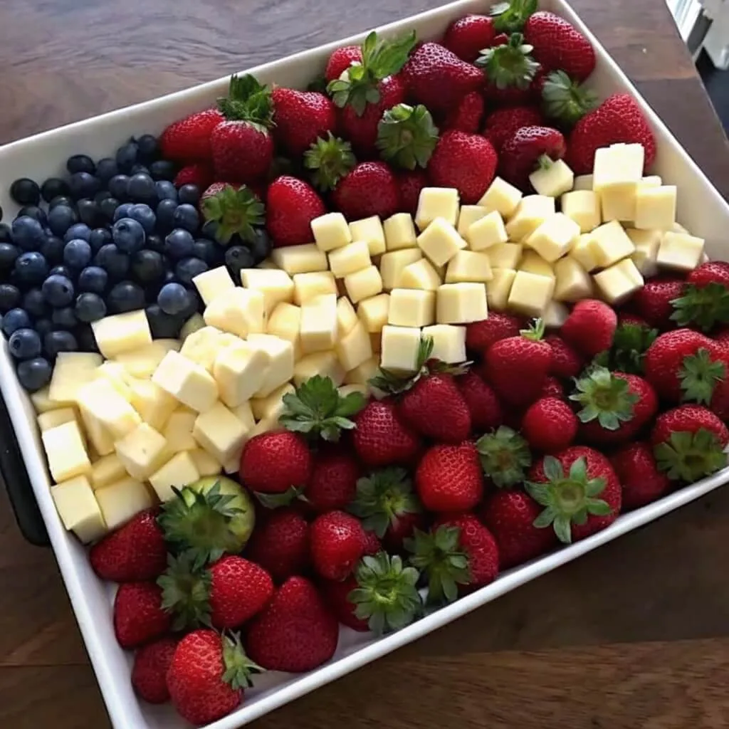 A deep white rectangular dish filled with blueberries, whole strawberries with green stems, and white cheese cubes in a flag arrangement.