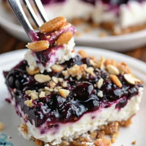 Close-up of a layered dessert bar with a graham cracker crust, thick vanilla cheesecake center, and a chunky blueberry topping sprinkled with crushed nuts.