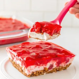 A pink spatula lifting a square piece of strawberry pretzel dessert over the main dish.