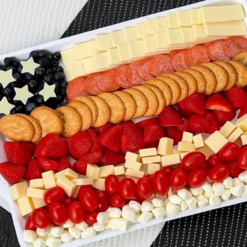 A 4th of July charcuterie board arranged like the American flag with rows of crackers, cheese, pepperoni, strawberries, tomatoes, and blueberries on a black and white geometric background.