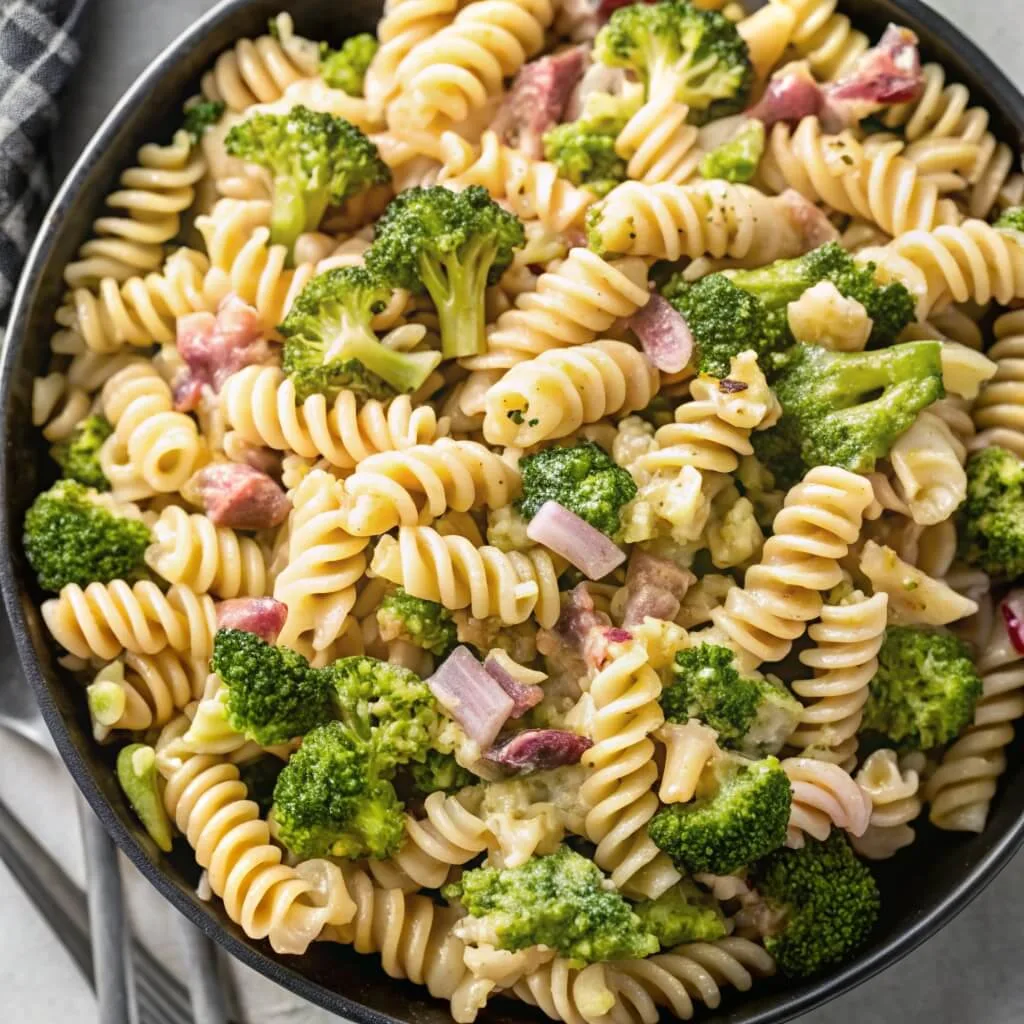 An overhead shot of a black bowl filled with rotini pasta salad, fresh broccoli florets, red onion, and pieces of savory bacon.