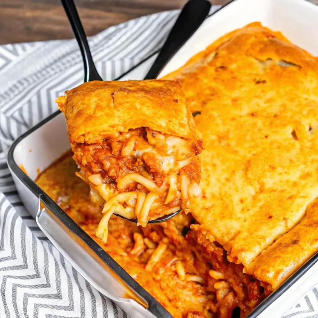 A black spatula lifting a large, cheesy slice of Million Dollar Spaghetti casserole from a white baking dish, showing layers of pasta and meat sauce.