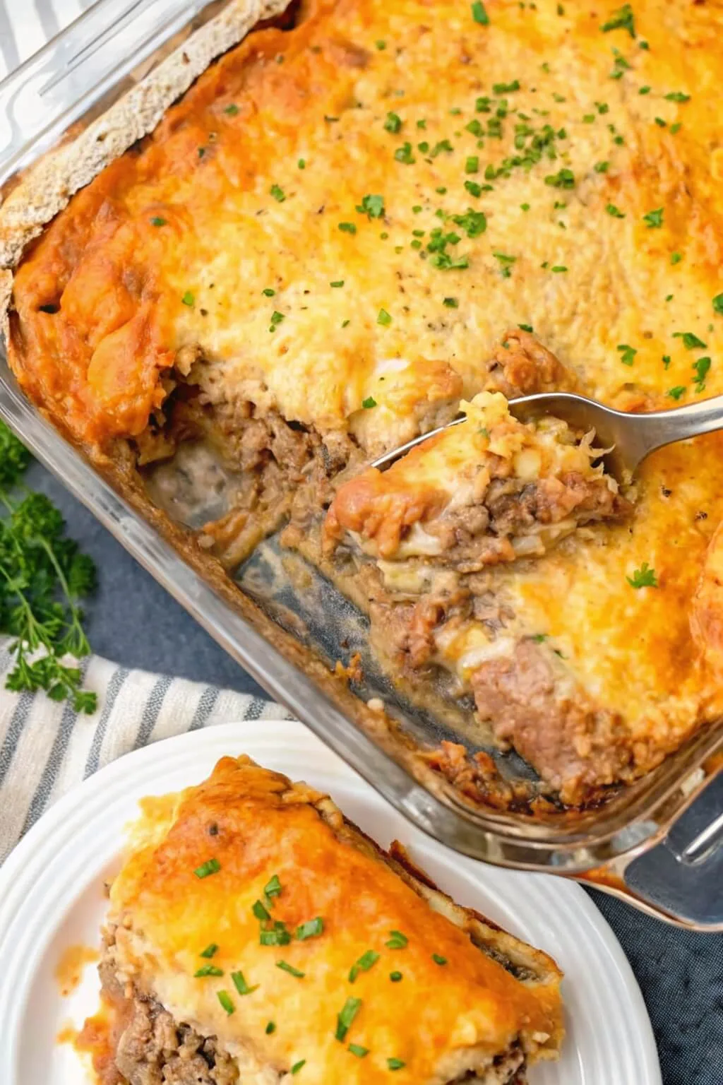 A glass baking dish filled with a layered ground beef and potato casserole, featuring a bubbly golden cheese topping and fresh parsley garnish.