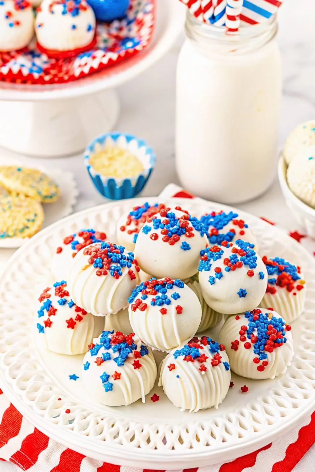 A white lattice-edged plate filled with white chocolate dipped cake pops decorated with red and blue star-shaped sprinkles, served with a bottle of milk.