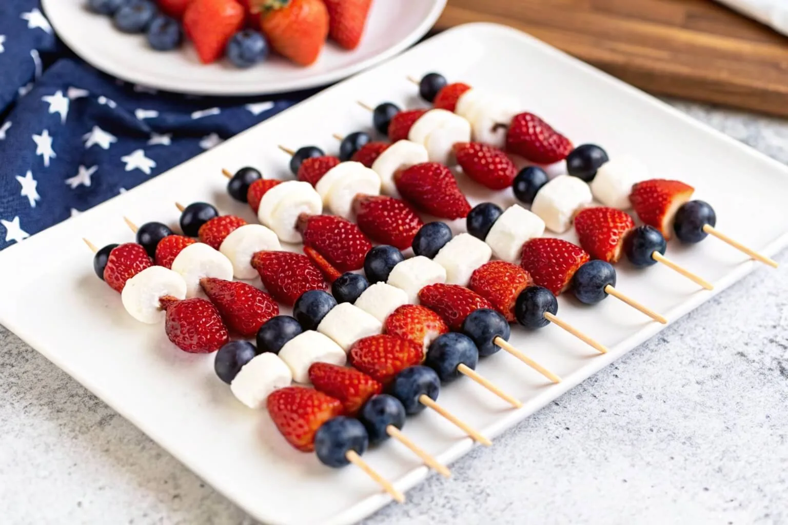 A white rectangular plate with five fruit skewers made of fresh strawberries, blueberries, and star-shaped white chocolate or cheese, set against a red and blue background.