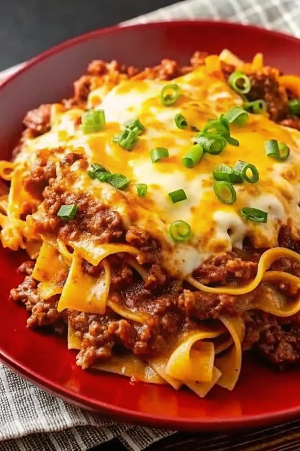 Plated beef goulash with wide egg noodles covered in melted cheddar cheese and chopped green onions.