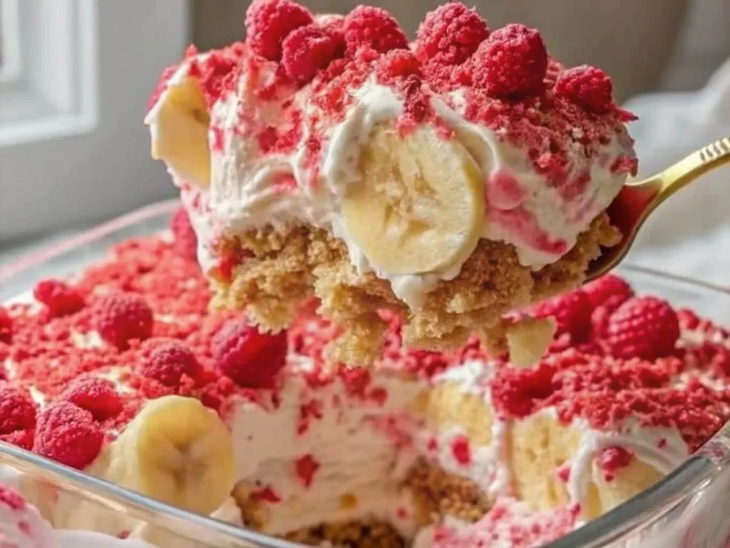 A golden spoon lifting a scoop of no bake raspberry banana icebox cake from a glass dish with whipped cream and fresh fruit.
