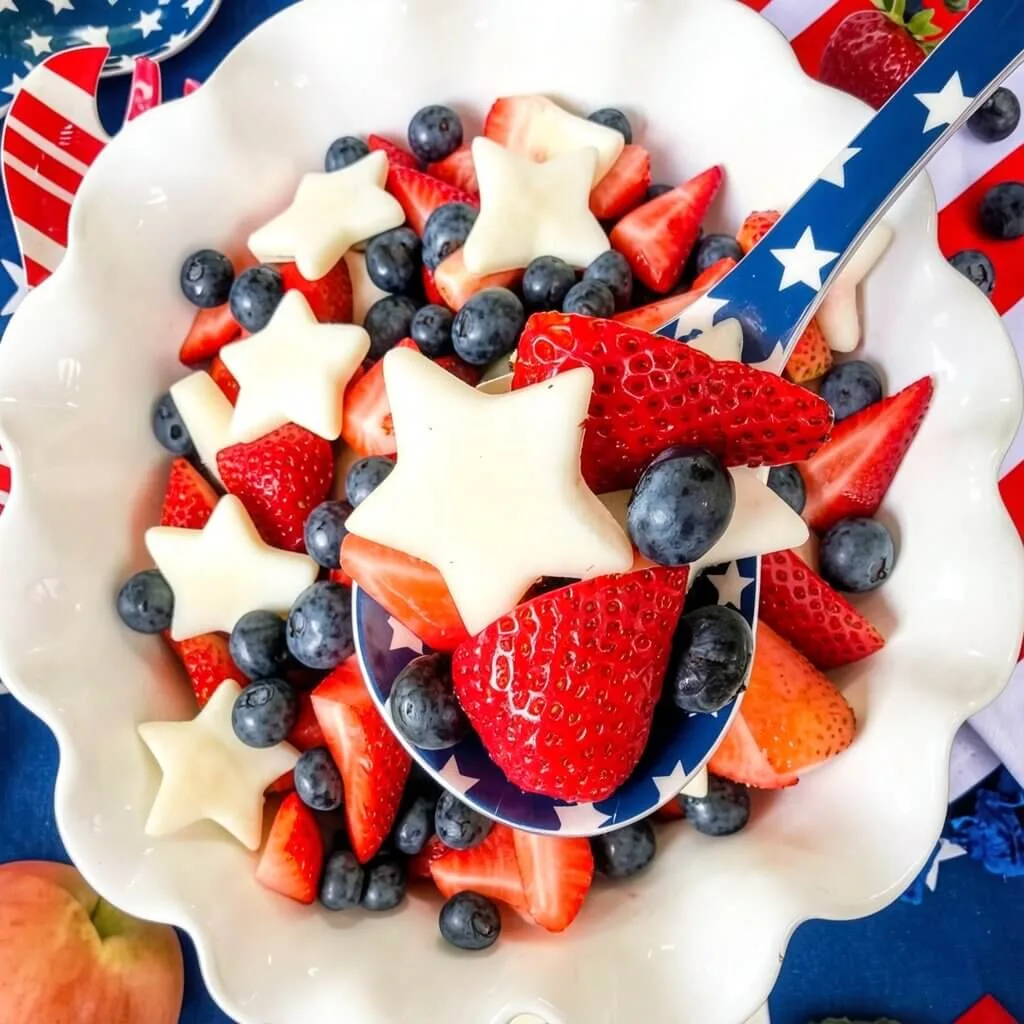 Overhead view of an easy 4th of July fruit salad with fresh strawberries, blueberries, and star-shaped apples being scooped with a blue star spoon.