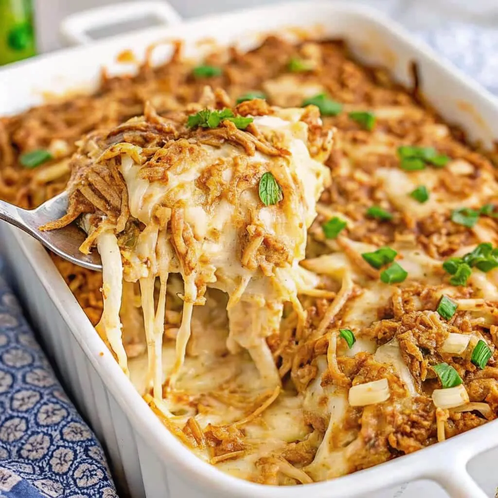 A close-up shot of a spoon lifting a cheesy serving of French onion chicken bake with crispy fried onions and fresh parsley from a white casserole dish.