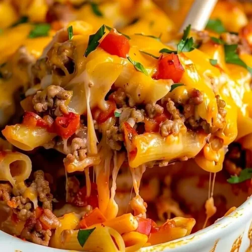 A wooden spoon lifting a scoop of cheesy taco pasta bake with ground beef, penne noodles, diced tomatoes, and melting cheddar cheese.