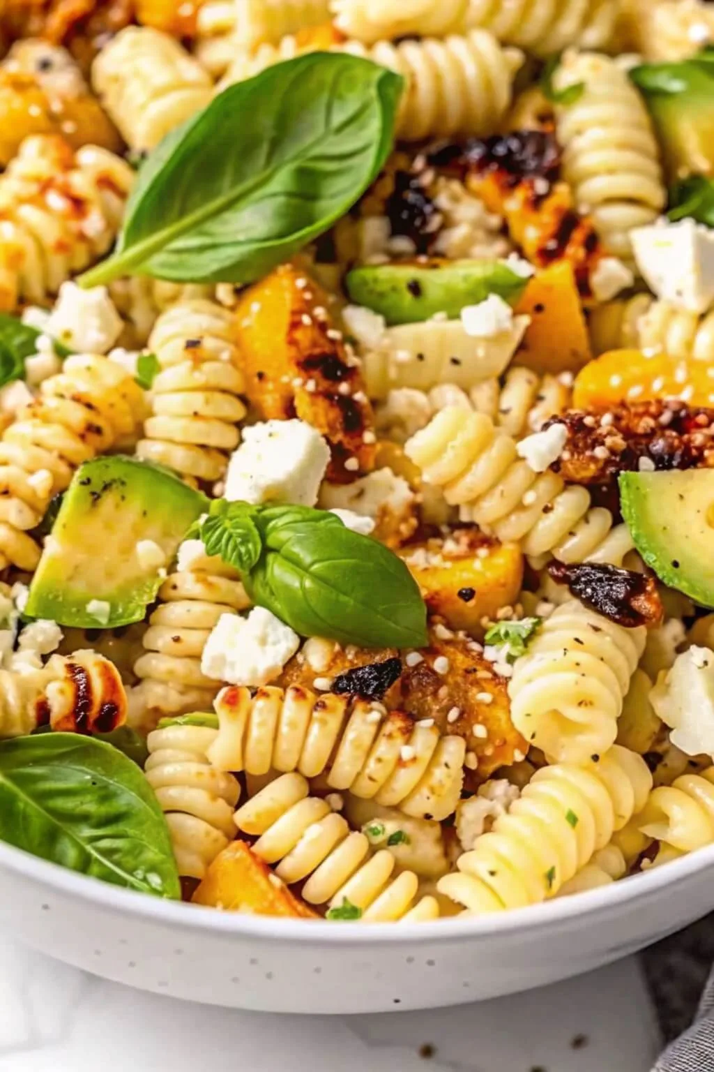 Close up of rotini pasta salad with grilled avocado chunks, crumbled feta cheese, and fresh basil leaves.