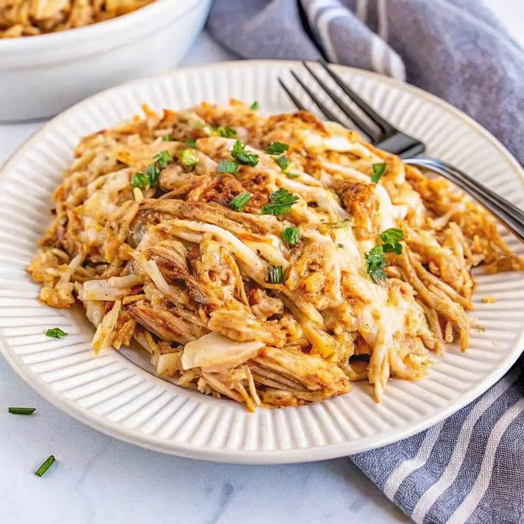 A serving of shredded French onion chicken bake on a white ribbed plate, garnished with fresh herbs and served with a silver fork.