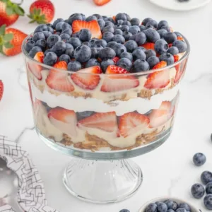 A large glass trifle bowl on a stand filled with layers of sponge cake, cream, sliced strawberries, and topped completely with fresh blueberries.