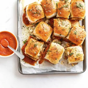 A baking sheet filled with a dozen baked meatball sliders topped with garlic herb butter and served with marinara dipping sauce.