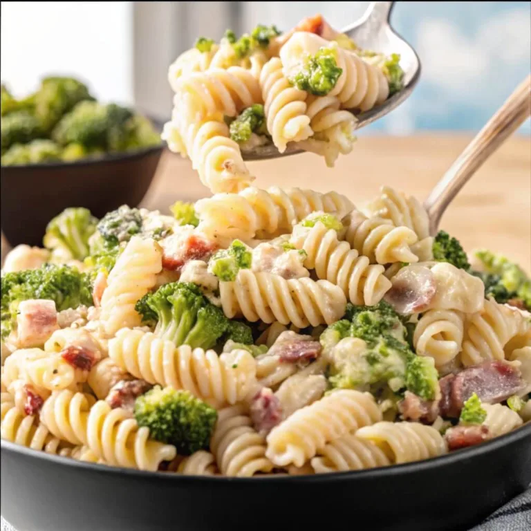 A silver spoon lifting a serving of creamy rotini pasta with broccoli and bacon from a large black bowl.