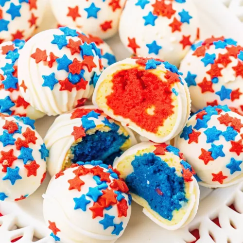 An overhead close-up of patriotic cake pops, with two halved to show the bright red and deep blue cake interiors inside the white chocolate shells.
