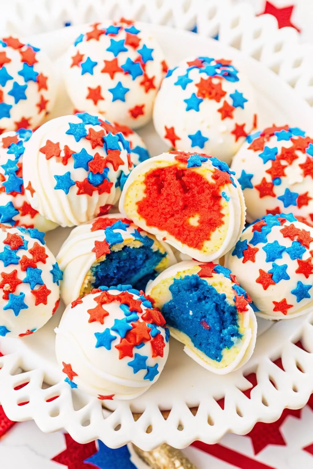 An overhead close-up of patriotic cake pops, with two halved to show the bright red and deep blue cake interiors inside the white chocolate shells.