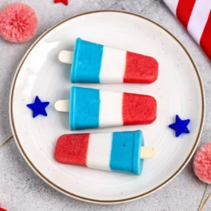 Three layered red, white, and blue popsicles arranged on a white plate with blue star decorations and pink pom-poms.