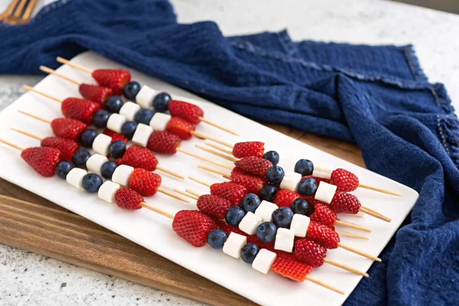 A close-up of red, white, and blue fruit skewers on a white plate, highlighting the fresh textures of the strawberries and blueberries.