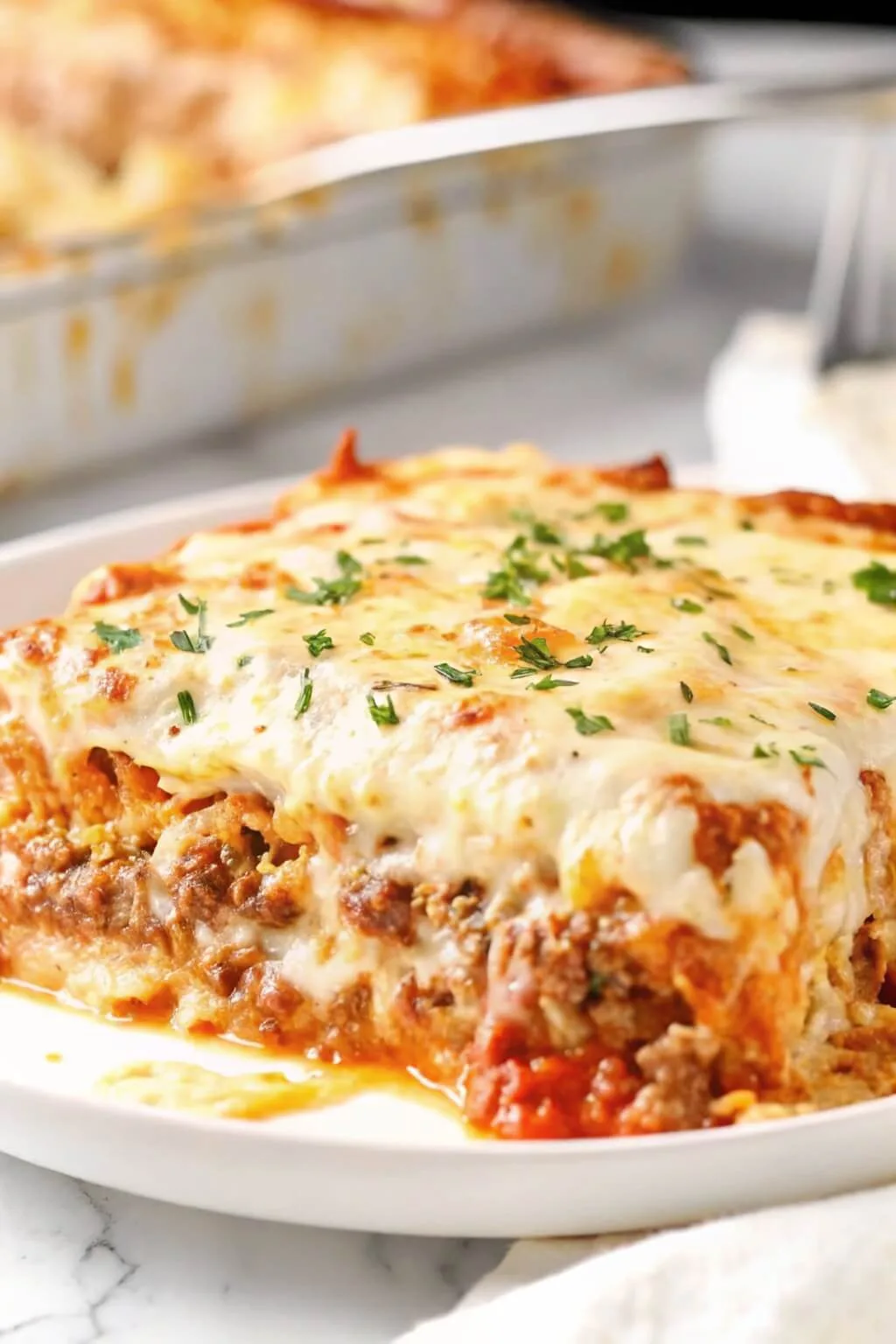 A thick square slice of meat lasagna topped with a bubbly golden cheese crust and fresh parsley on a plate.