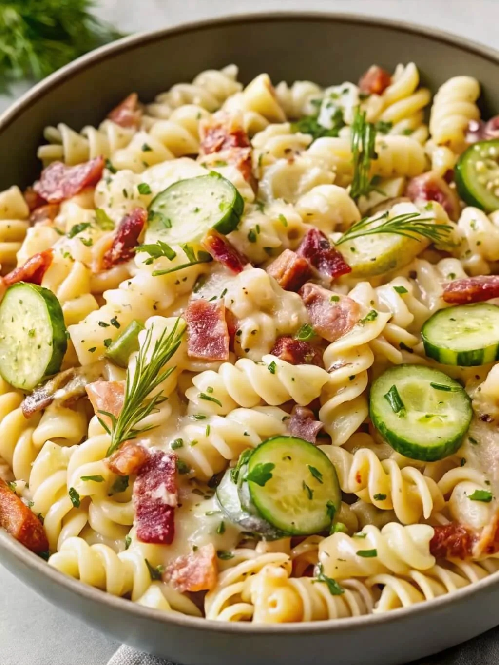 Creamy rotini pasta salad with crisp cucumber slices, smoky bacon pieces, and fresh herbs in a grey bowl.