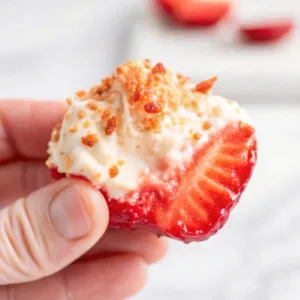 A hand holding a single cream cheese stuffed strawberry bite topped with crushed graham cracker crumbs.