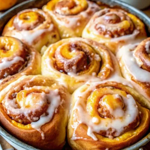 An overhead view of a round baking pan filled with freshly baked, glazed peach sweet rolls next to fresh peaches.