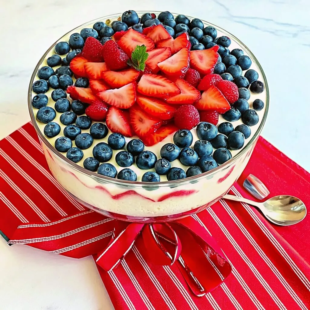 A glass trifle bowl with a red ribbon at the base, topped with a dense pile of sliced strawberries and a ring of blueberries.