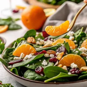 A gold fork lifting a single juicy mandarin orange segment from a bowl of fresh spinach and cranberry salad.