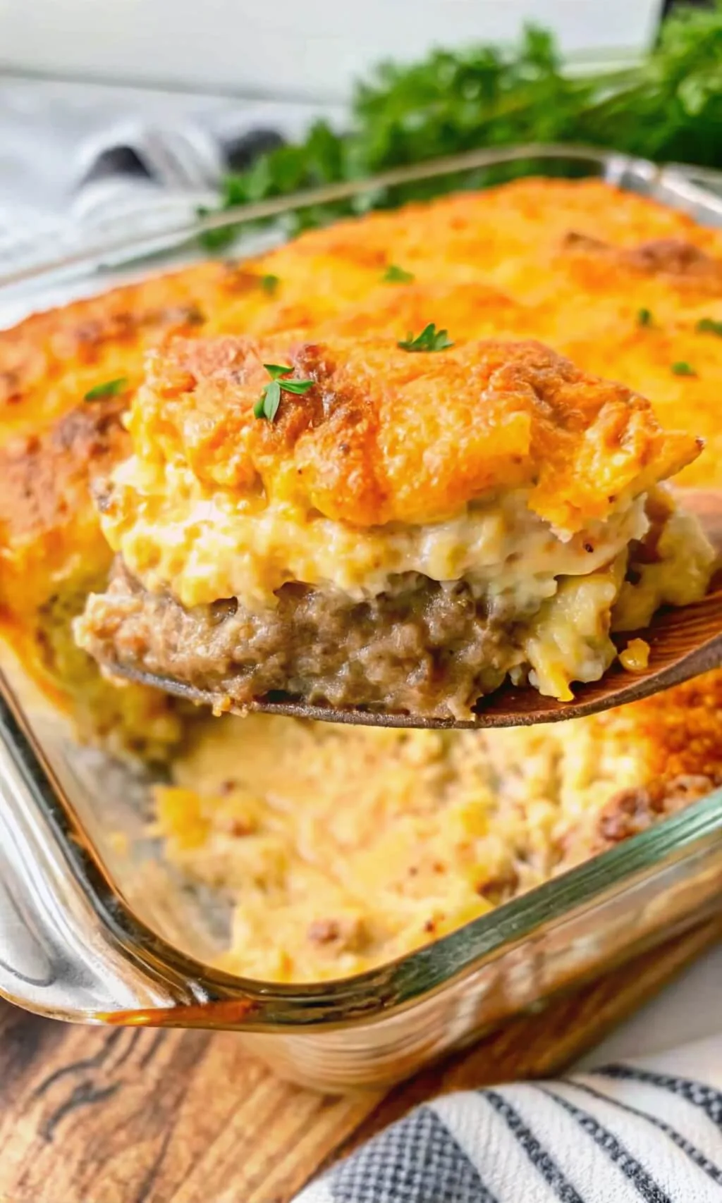 A wooden spoon lifting a large square slice of a three-layer meat and potato casserole from a glass pan.