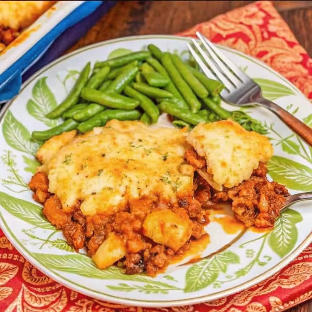 A decorative plate featuring a large slice of Shepherd's Pie with a bubbly cheese crust, served with a side of steamed green beans on a floral patterned plate.