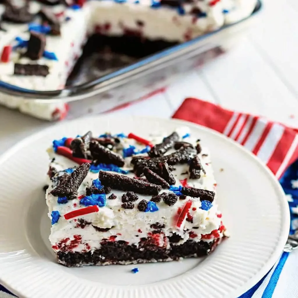 A single square slice of patriotic red velvet cake on a white plate, topped with crushed Oreos and blue and red sprinkles.