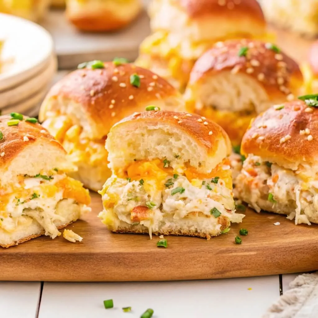 A wooden board filled with toasted brioche sliders stuffed with creamy chicken salad, melted cheese, and green onion garnish.