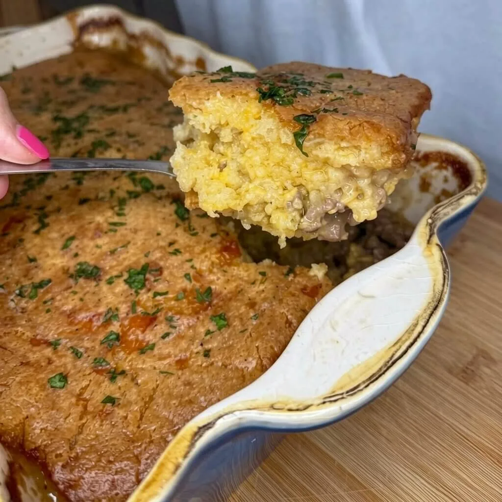 A metal spatula lifting a slice of easy cheeseburger cornbread casserole from a blue baking dish, showing a seasoned ground beef and yellow cornbread filling.
