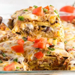 A metal spatula lifting a slice of easy Mexican lasagna from a glass baking dish, showing layers of tortillas, ground beef, black beans, and melted cheese.