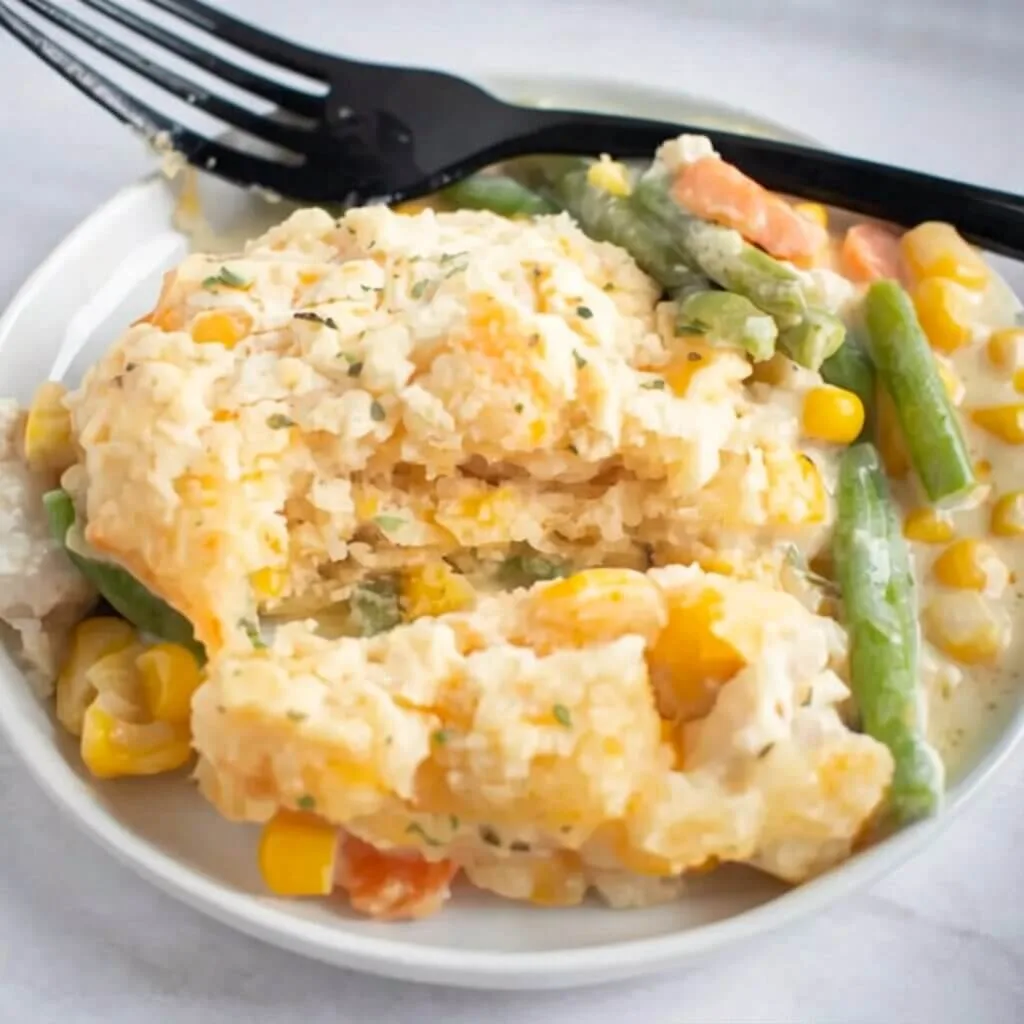A close up individual serving of an easy chicken pot pie biscuit casserole on a white plate with green beans, sweet corn, and carrots.