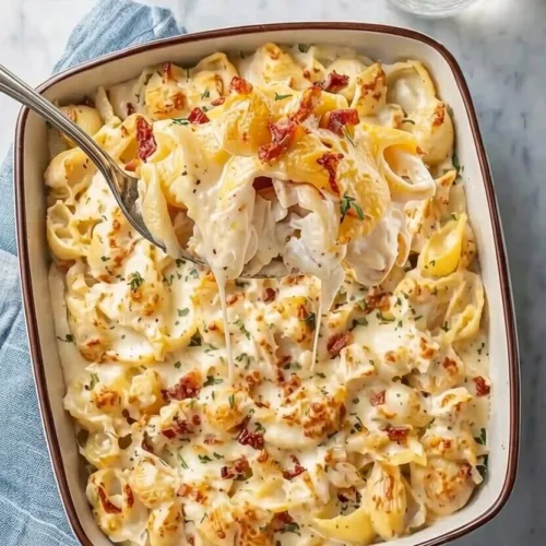 Overhead top down view of an easy garlic chicken pasta bake in a ceramic dish, with a large spoon scooping up cheesy pasta shells topped with crispy bacon.