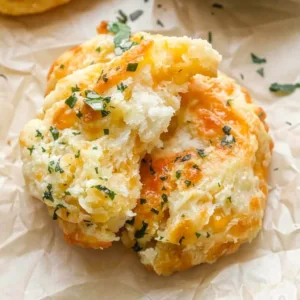 A close up shot of an easy cheddar bay biscuit broken open on parchment paper, showing a fluffy interior, melted cheddar cheese, and garlic herb butter.