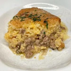 A single serving of ground beef tamale pie recipe on a white textured plate showing distinct layers of seasoned meat and sweet cornbread.