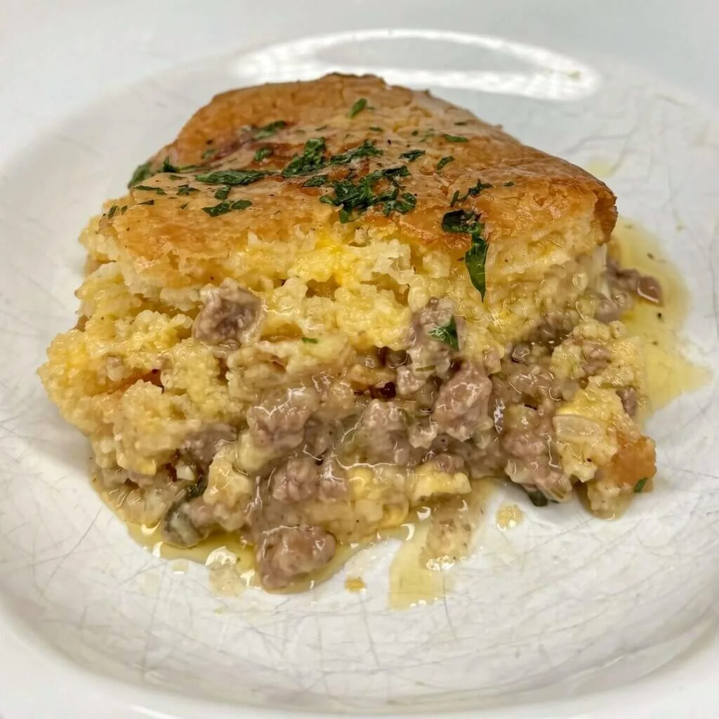 A single serving of ground beef tamale pie recipe on a white textured plate showing distinct layers of seasoned meat and sweet cornbread.